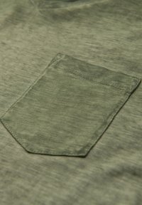 Tkanina olivově zelené košile s viditelnou kapsou. Textura vypadá jemně s mírně opotřebovaným vzhledem. Kapsa má obdélníkový tvar a čisté prošívání.