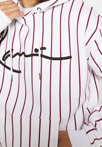 Felpa con cappuccio bianca con strisce verticali rosse e nere, dotata di logo ricamato nero, coulisse regolabile e tasca frontale.