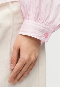 Μανσέτα ροζ ανοιχτού χρώματος με υφή, featuring a gathered design και ένα μόνο ροζ κουμπί, συνδυασμένη με ύφασμα σε κρεμ χρώμα από κάτω.