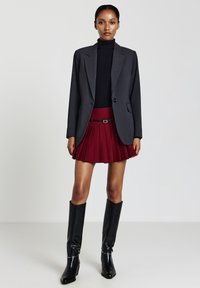 Donna che indossa un blazer grigio scuro, un dolcevita nero, una gonna mini plissettata rossa con cintura e stivali neri al ginocchio, in piedi di fronte a uno sfondo semplice.