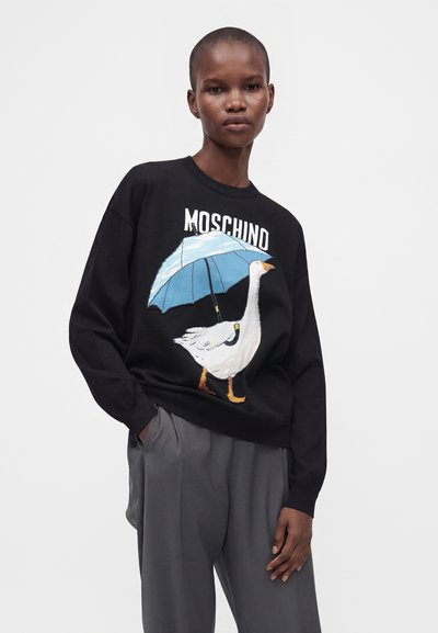 Άτομο που φοράει μαύρο πουλόβερ Moschino με σχέδιο μιας λευκής χήνας που κρατά μια μπλε ομπρέλα, σε συνδυασμό με γκρι παντελόνια.