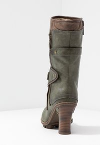 Bota de tobillo de cuero verde con un puño marrón y detalles de cremallera, que destaca una correa decorativa y un tacón grueso sobre un fondo blanco liso.