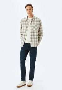 Camisa a cuadros en beige con líneas azules y marrones; bolsillo frontal; combinada con unos vaqueros azul oscuro y zapatillas crema con suelas marrones.