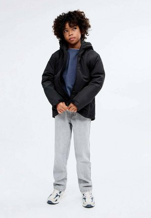 Veste noire matelassée avec capuche, portée ouverte sur un sweat-shirt bleu. Associée à un jean gris et des baskets blanches avec des accents navy.