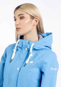 Chaqueta de color azul claro hecha de un tejido suave. Cuenta con capucha, cordones, botones a presión y un logotipo blanco en el pecho. Textura suave.