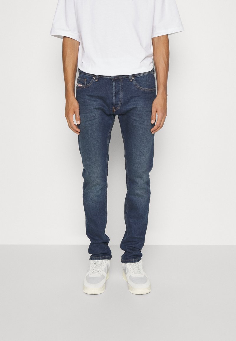 Homme portant un jean slim bleu foncé, des baskets blanches et une chemise blanche unie à manches courtes, debout sur un sol gris clair.
