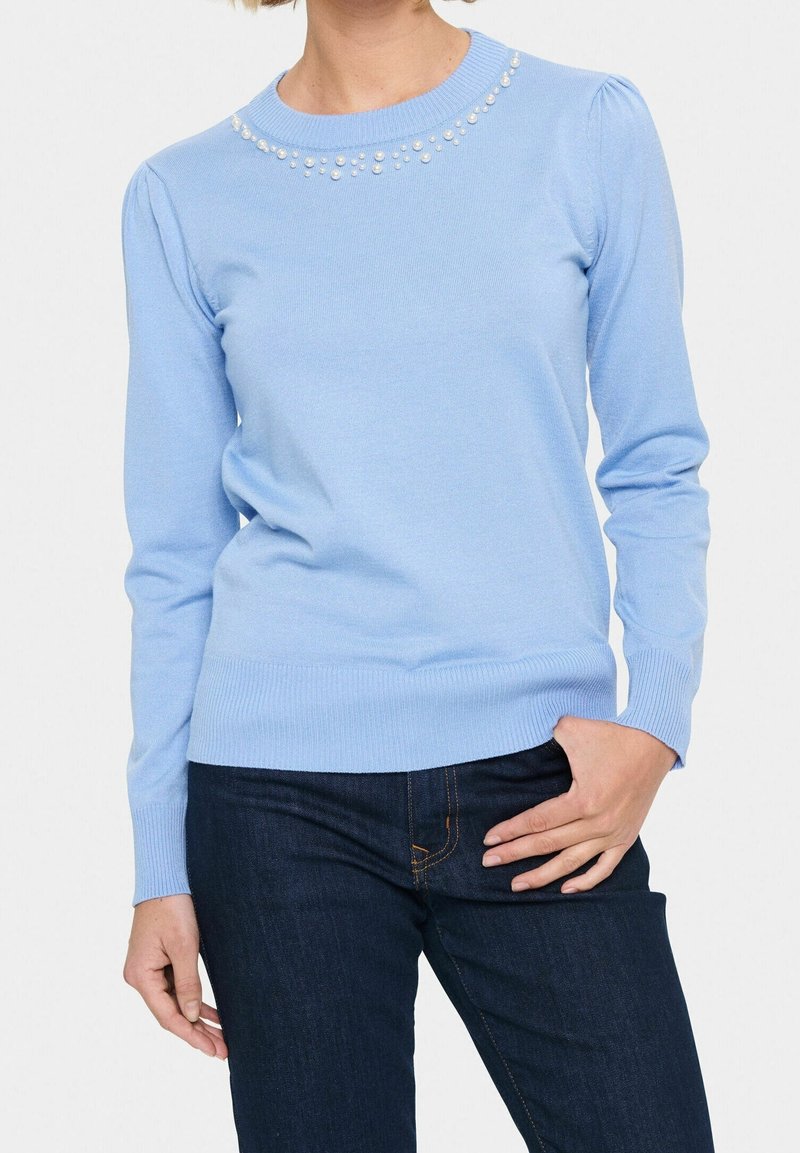 Femme portant un pull à manches longues bleu clair avec des détails en perles près de l'encolure et un jean bleu foncé, la main posée sur la hanche.