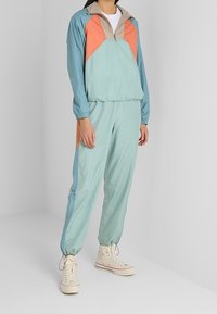 Lättvikts, färgblockerat träningsset med en mintgrön, orange och beige design; har en jacka med halvdragkedja samt elastiska ärmslut och anklar.