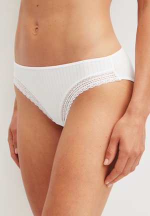 Donna che indossa biancheria intima bianca a costine con bordo in pizzo, vista dalla zona dell'addome fino alla coscia superiore su sfondo neutro.