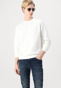 Sudadera blanca con cuello redondo y puños de canalé, combinada con jeans de mezclilla oscura. El modelo lleva gafas de sol redondas. Diseño sencillo, ajuste casual.