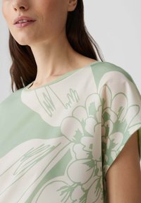 Blusa verde chiaro con motivi floreali in bianco e beige. Presenta un'ampia scollatura e un tessuto morbido e liscio con una vestibilità comoda.