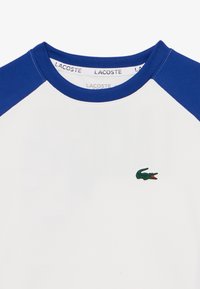 T-shirt blanc à manches courtes avec des manches raglan bleues, arborant un petit logo de crocodile vert à l'avant et un col rond classique.