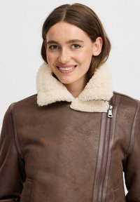 Veste en cuir marron avec un col en shearling, présentant une fermeture zippée et une finition texturée. Des poches fonctionnelles complètent le design.