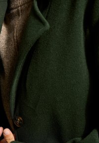 Käsi, joka holds a dark green wool coat (villako) avattuna, joka paljastaa ruskean neuleen (neulepuseron) alla, keskittyen kankaan tekstuureihin ja nappiyksityiskohtiin.
