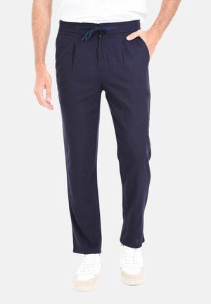 Pantaloni blu navy con vita regolabile, pieghe davanti e tasche laterali; abbinati a sneaker bianche su uno sfondo neutro.