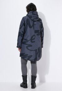 Cappotto con cappuccio, impermeabile, di colore grigio scuro con motivi astratti neri. Presenta un orlo posteriore più lungo e tasche laterali, abbinato a leggings grigi e stivali neri.