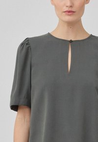 Kurzärmelige, dunkelgraue Bluse mit einem runden Ausschnitt, die eine kleine Schlüsselloch-Öffnung und einen dekorativen Knopf am Hals hat. Glatter Stoff.