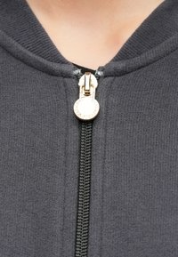 Ciemnoszara bluza z kapturem zapinana na zamek, wykonana z fakturowanego materiału, z czarnym zamkiem błyskawicznym oraz złotym suwakiem z logo przy kołnierzu.