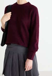 Pull en laine bordeaux avec un col rond et des poignets côtelés, associé à une jupe plissée grise. Détail d'un sac à main noir inclus.