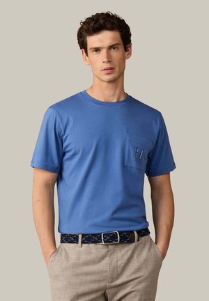 Joven con camisa azul de manga corta con bolsillo en el pecho y pantalones beige, de pie con las manos en los bolsillos frente a un fondo liso.