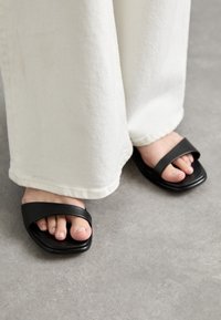 Sandales noires à glissière avec deux larges brides, design à bout carré, texture lisse, portées avec un pantalon blanc ample.