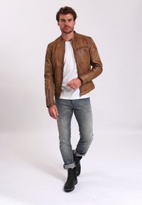 Giacca in pelle marrone con finitura texturizzata, tasca con zip e colletto alto, abbinata a jeans azzurri chiari e stivali neri alla caviglia.