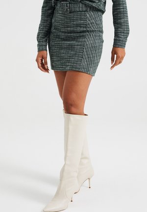 Dopasowana zielona mini spódnica w teksturę z wzorem w kratę i paskiem w talii, zestawiona z białymi kozakami do kolan ze skóry.