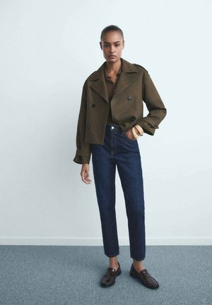 Olivovo zelená skrátená dvojradová bunda, hnedý rebrovaný top, vysoké tmavé džínsy a tmavé mokasíny, s kruhovým náramkom na ľavom zápästí.