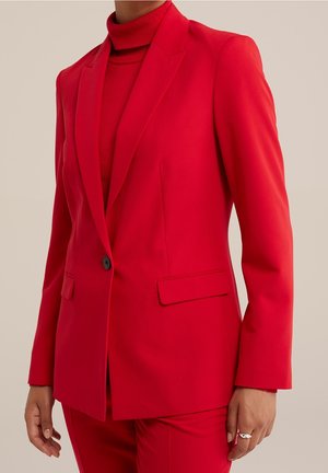 Persoon draagt een nauwsluitend felrood blazer met een enkele zwarte knoop over een bijpassende rode coltrui en rode broek.