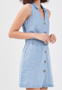 Robe en denim sans manches de couleur bleu clair, ornée de boutons en écaille de tortue sur le devant et agrémentée d'une ceinture assortie avec boucle ronde, portée par une personne ayant des cheveux bouclés.