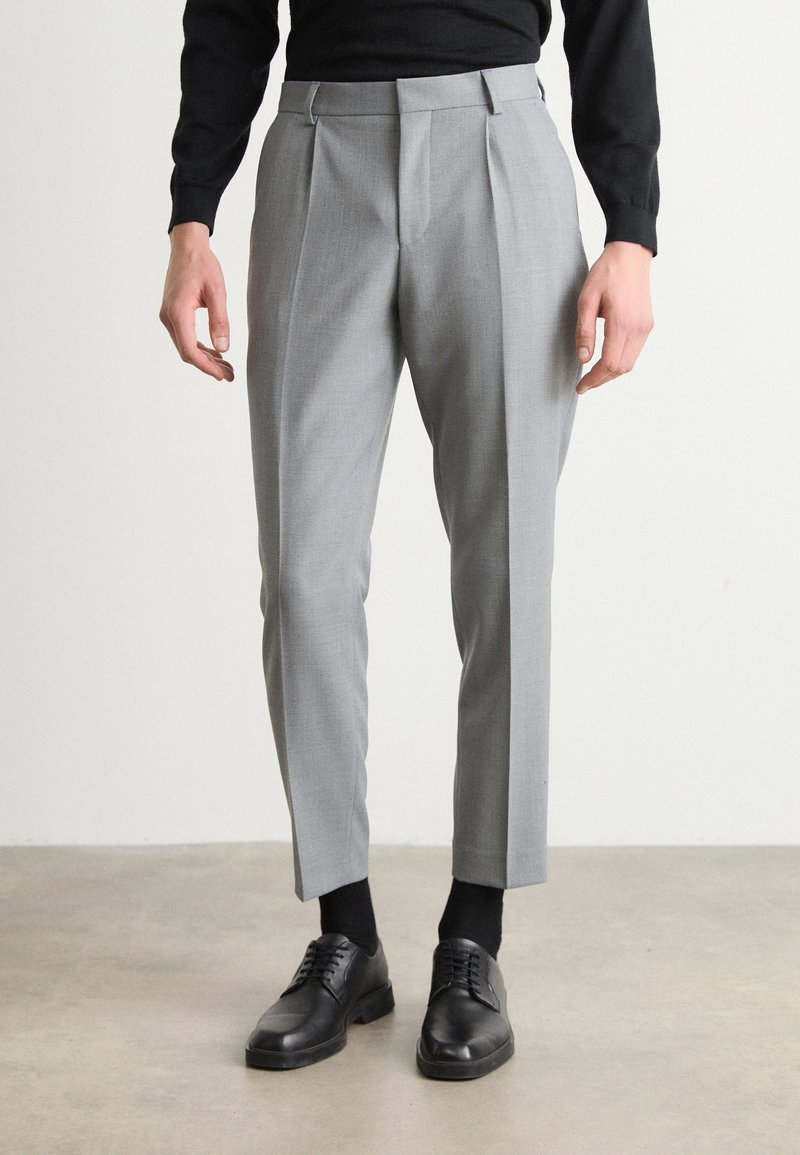 Gray pleated trousers with a tailored fit, fabric texture, side pockets, and a flat front design, paired with black shoes and socks.