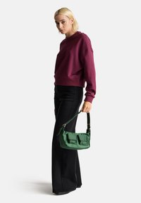 Bolso de hombro verde y elegante con dos bolsillos frontales, hecho de un material suave. Combinado con un sudadera ajustada burdeos y pantalones acampanados negros.