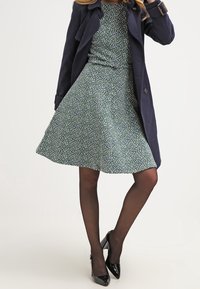 Vihreä kukkamekko, jossa on istuva yläosa ja levenevä hame, yhdistettynä laivastonsiniseen trench-coatiin. Asusteena mustat korkokengät ja läpinäkyvät mustat sukkahousut.