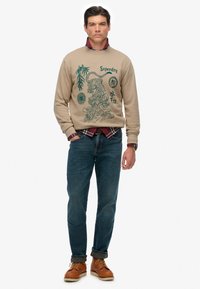 Uomo che indossa un felpa beige con stampa di tigre verde e testo, una camicia a quadri rossa sotto, jeans blu e stivali beige, in piedi con una mano in tasca.