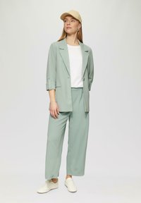 Helles grünes Blazer mit hochgekrempelten Ärmeln, passenden weiten Hosen und einem weißen T-Shirt. Eine beigen Kappe und weiße Sneaker runden den Look ab.