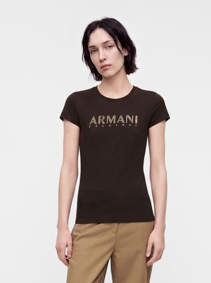 Ung person med kort mørkt hår iført en tætsiddende sort Armani Exchange t-shirt og beige bukser, stående mod en ensfarvet lys baggrund.