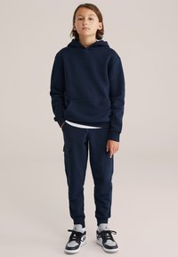 Marineblauwe hoodie met kangoeroezak en bijpassende joggers, met omslagmanchetten en cargopockets. Gemaakt met zwarte en grijze sneakers.