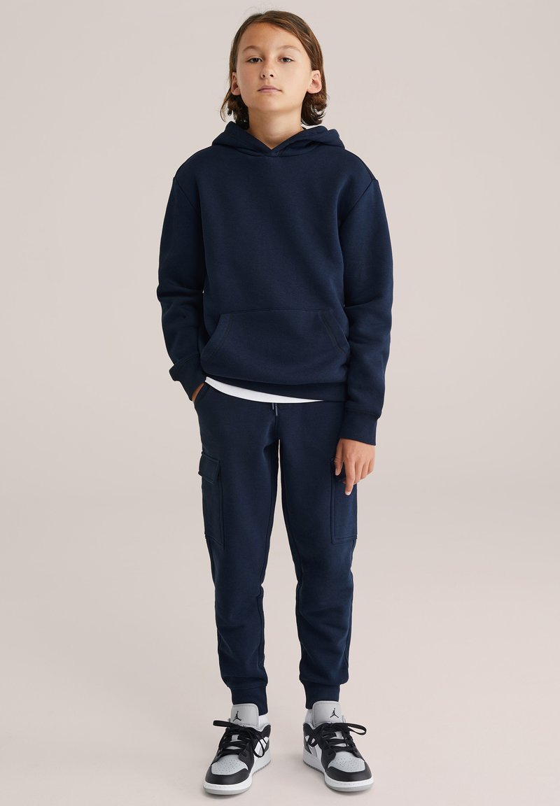 Marineblauwe hoodie met kangoeroezak en bijpassende joggers, met omslagmanchetten en cargopockets. Gemaakt met zwarte en grijze sneakers.