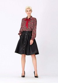 Blouse florale rouge avec un nœud, associée à une jupe noire évasée avec un fermoir à boutons à l'avant et des poches. Le mannequin porte des talons noirs.