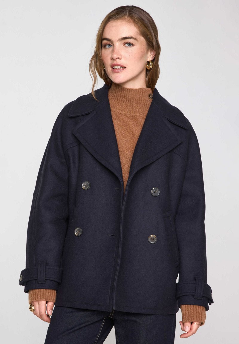 Navy wollen-blend dubbele-breasted jas met grote revers en twee voorkruiszakken. Voorzien van zilverkleurige knopen en brede mouwen over een bruine trui.