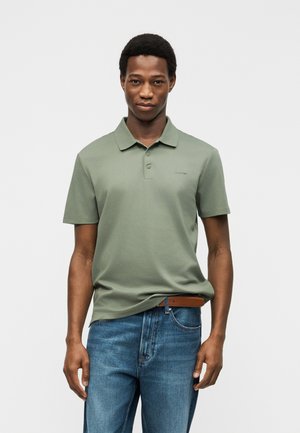 Άνδρας με πράσινο κοντομάνικο πόλο και μπλε τζιν, στέκεται μπροστά σε απλό φωτεινό φόντο, κοιτάζοντας μπροστά με ουδέτερη έκφραση.