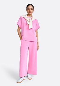 Roze katoenmengsel oversized top en broek, met wijde pijpen en opgerolde hemden, gecombineerd met witte sneakers. Lichte gebreide trui om de nek gedrapeerd.