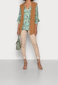 Mujer vistiendo pantalones beige, blusa floral verde, chaleco sin mangas marrón, tacones beige, sosteniendo un bolso verde frente a un fondo blanco.