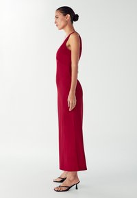 Červené priliehavé šaty bez rukávov, dlhý strih, hladká textúra, bočný rozparok, spárované s čiernymi sandálmi na remienky.