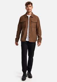Braune Wildlederjacke mit Knopfverschluss, zwei Brusttaschen und schwarzen Knöpfen, kombiniert mit einem weißen T-Shirt, schwarzen Hosen und schwarzen Stiefeln.