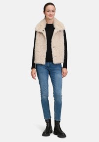 Gilet en fausse fourrure beige clair avec un large col et une fermoir à boutons. Porté sur une chemise noire à manches longues, associé à un jean bleu et des bottes noires.