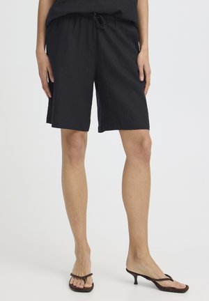 Kobieta w luźnych czarnych szortach do kolan z elastycznym pasem ze ściągaczem oraz brązowych niskich sandałach na cienkim pasku.