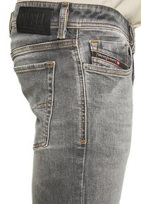 Jeans de mezclilla gris con un ajuste ceñido, con costuras amarillas en contraste, un parche de cuero negro en la cinturilla y dos bolsillos traseros.