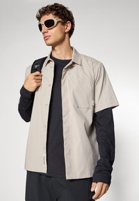 Helles beige Kurzarm-Hemd mit Knopfleiste über einem dunklen Langarm-Oberteil, mit einer Brusttasche und einem Etikett. Kombiniert mit beige Sonnenbrille.
