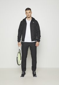 Čierna športová bunda s kapucňou, skombinovaná s bielym tričkom a čiernymi nohavicami. Model drží tenisovú raketu. Čierne tenisky dopĺňajú outfit.